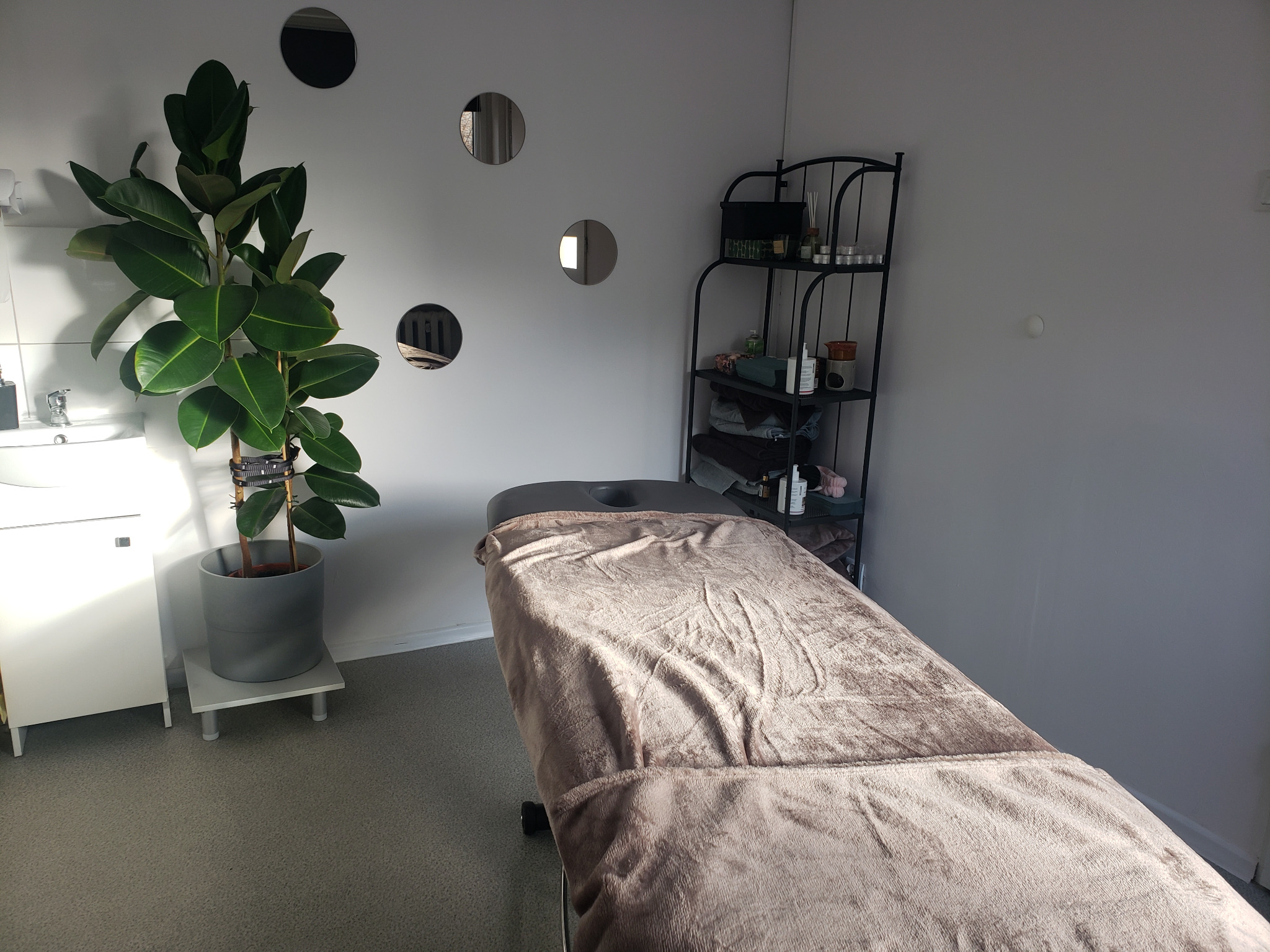 Klimatyczne wnętrze gabinetu z łóżkiem do zabiegów przykrytym pluszowym kocem, regałem z kosmetykami i dużym kwiatem doniczkowym obok umywalki.
