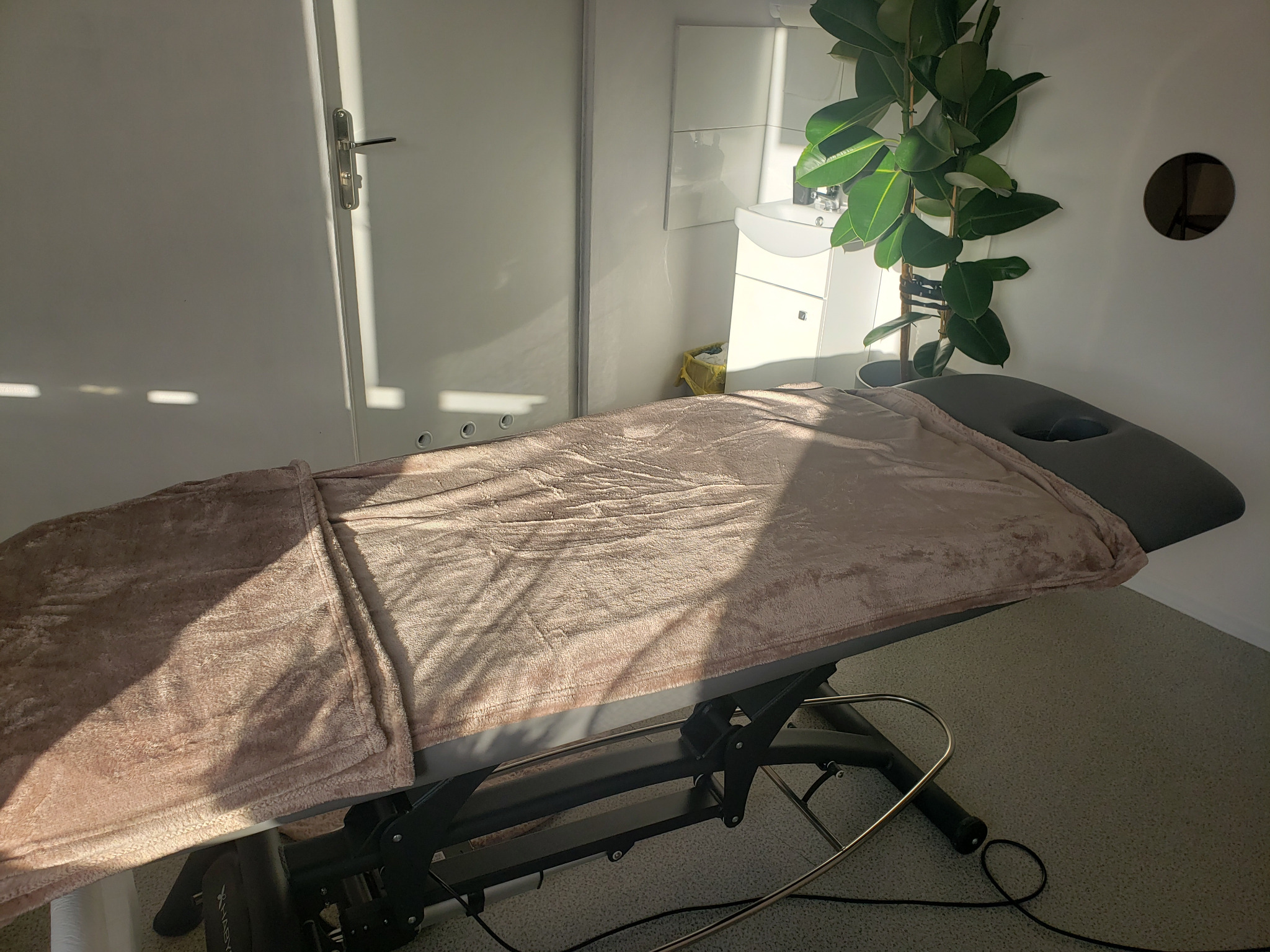 Regulowane łóżko do zabiegów z szarym zagłówkiem i różowym, pluszowym okryciem, oświetlone promieniami słonecznymi w minimalistycznym gabinecie z rośliną doniczkową.