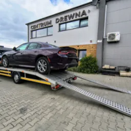 Laweta z fioletowym Dodge Charger na tle budynku 'Centrum Drewna'. Rampy lawety opuszczone, gotowe do zjazdu pojazdu. Pochmurne niebo.