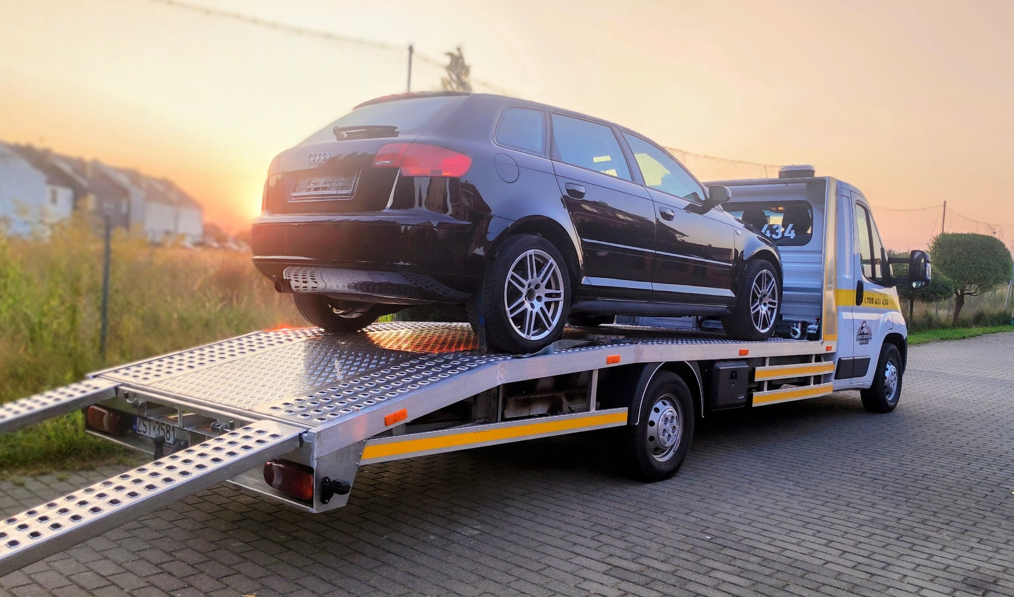 Czarny samochód Audi kombi na platformie lawety, z widocznym zachodem słońca w tle i numerem telefonu na kabinie pojazdu.