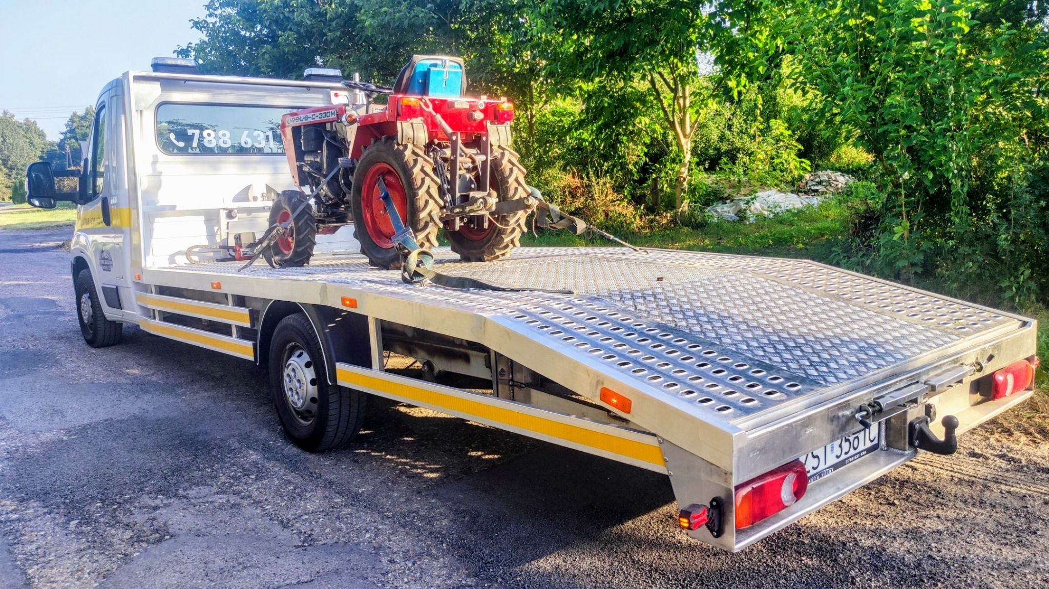 Czerwony traktor Ursus C-330M zabezpieczony pasami na platformie lawety, widok z tyłu, w tle zieleń.