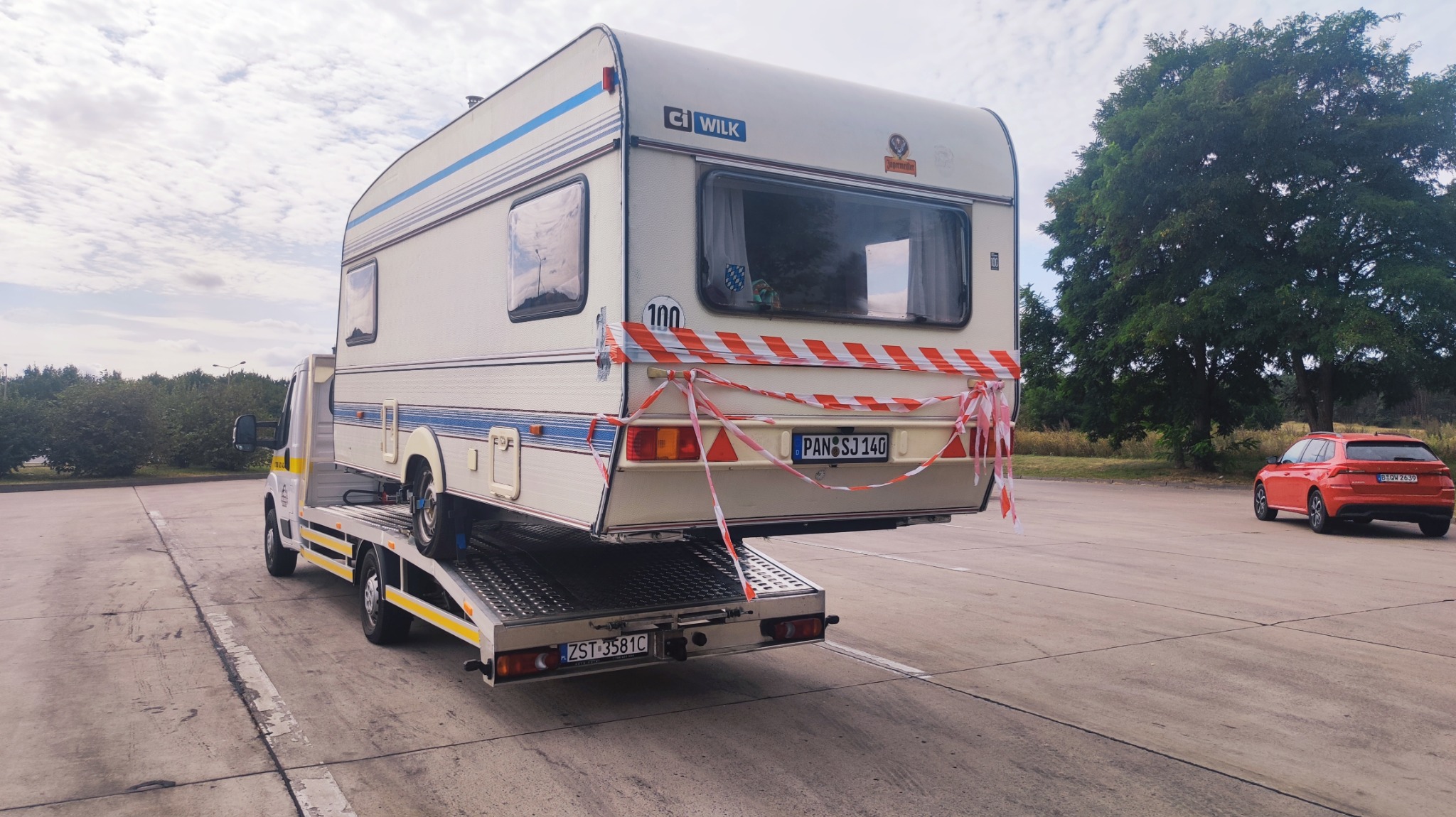 Przyczepa kempingowa marki Wilk zabezpieczona taśmą ostrzegawczą, przewożona na platformie niskopodwoziowej. W tle widoczny czerwony samochód osobowy i drzewa.
