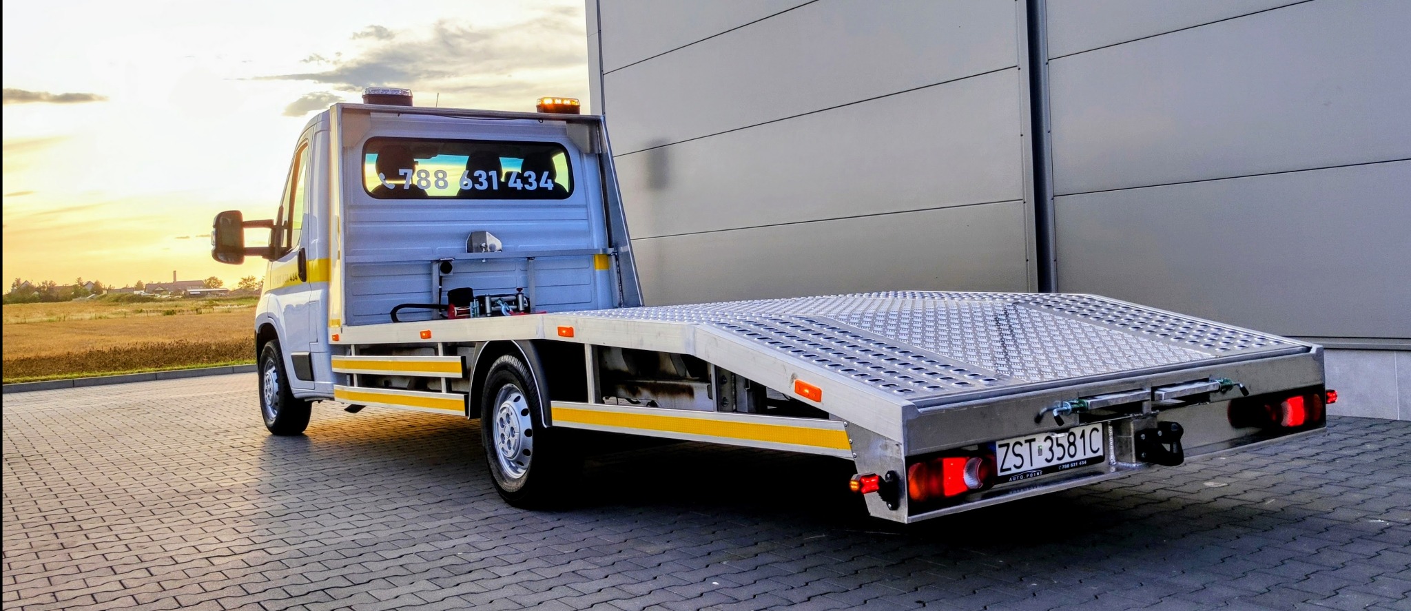 Nowa laweta z aluminiową platformą, widok z tyłu, z numerem telefonu na szybie kabiny, zaparkowana na kostce brukowej, z polem w tle i budynkiem przemysłowym po prawej stronie, w promieniach...