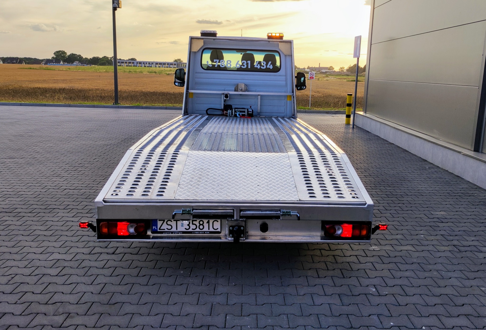 Tył srebrnej lawety z widoczną tablicą rejestracyjną ZST 3581C na tle pola i budynku, z numerem telefonu 788 631 434 w oknie kabiny.