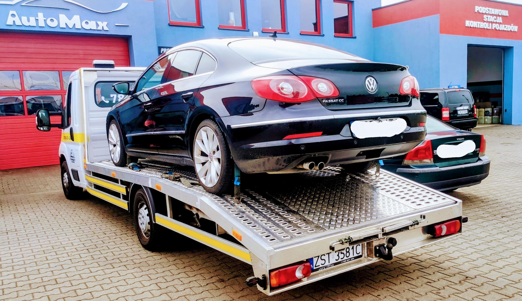 Czarny Volkswagen Passat CC zabezpieczony pasami na platformie lawety, z widocznym budynkiem stacji kontroli pojazdów w tle.