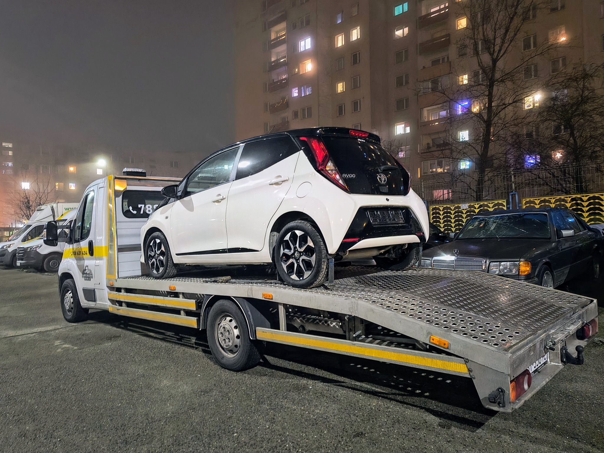 Biały samochód osobowy Toyota Aygo z czarnym dachem, przewożony na platformie lawety w nocy, na tle bloku mieszkalnego z oświetlonymi oknami.