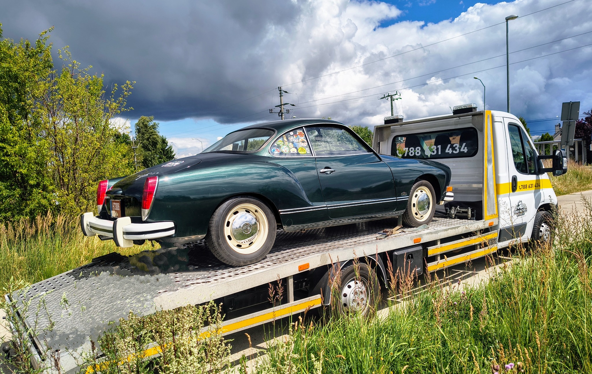 Zielony, zabytkowy Volkswagen Karmann Ghia na lawecie z numerem telefonu, na tle pochmurnego nieba i zieleni. Transport specjalistyczny.