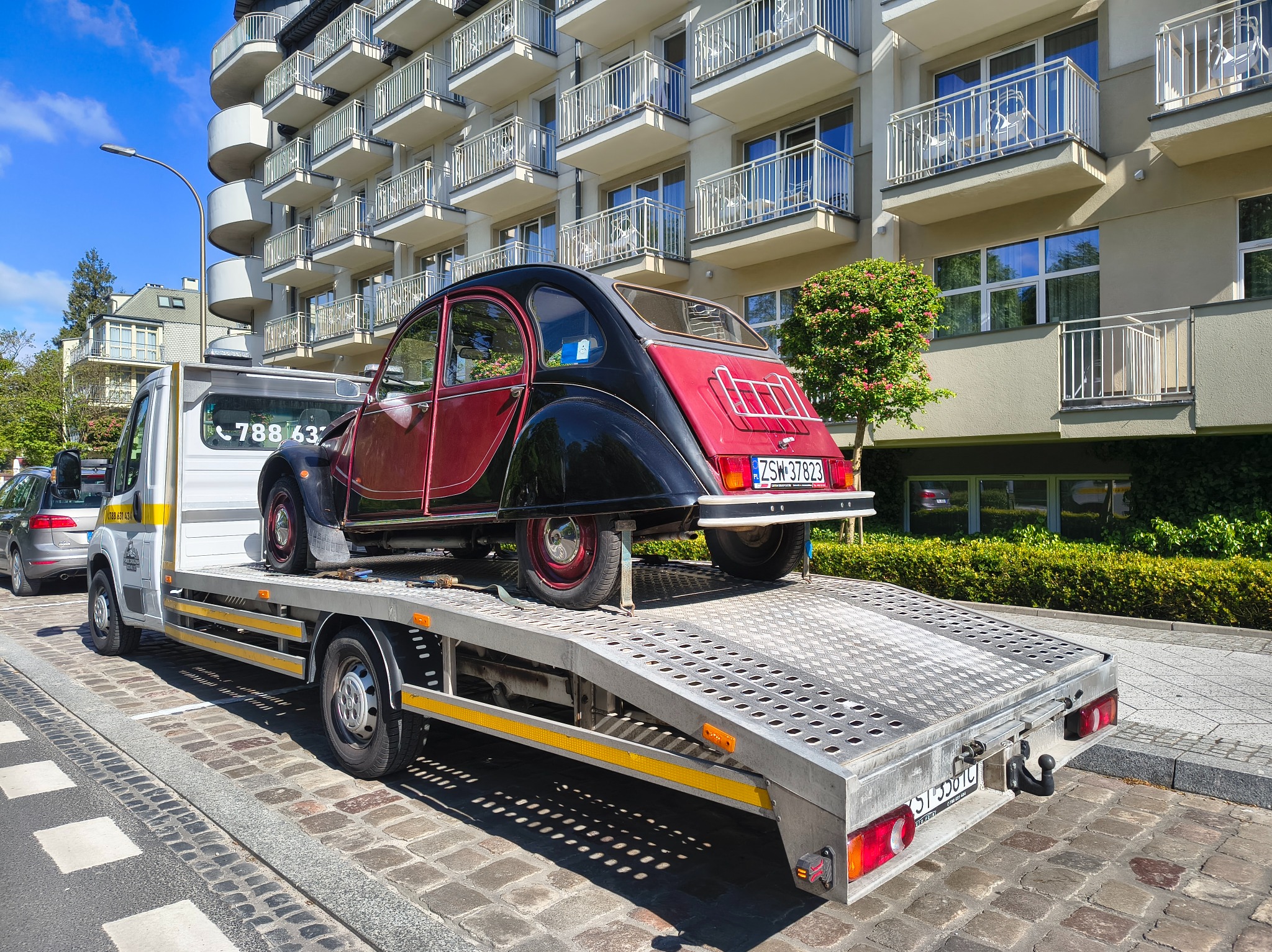 Laweta przewozi czerwono-czarne, zabytkowe auto na tle budynku z balkonami. Tablica rejestracyjna pojazdu ZSW 37823. Dzień słoneczny.