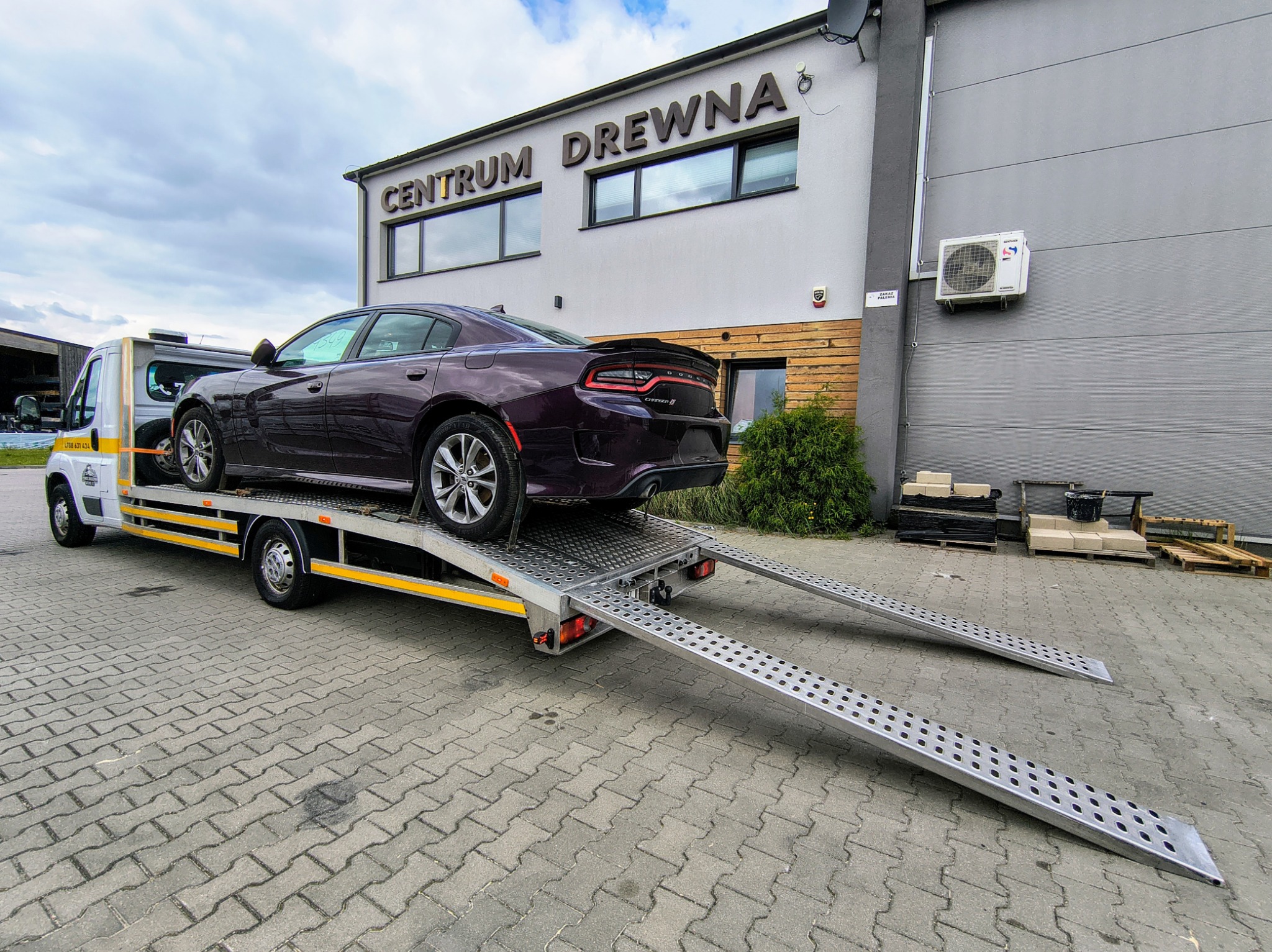 Laweta z fioletowym Dodge Charger na tle budynku 'Centrum Drewna'. Rampy lawety opuszczone, gotowe do zjazdu pojazdu. Pochmurne niebo.