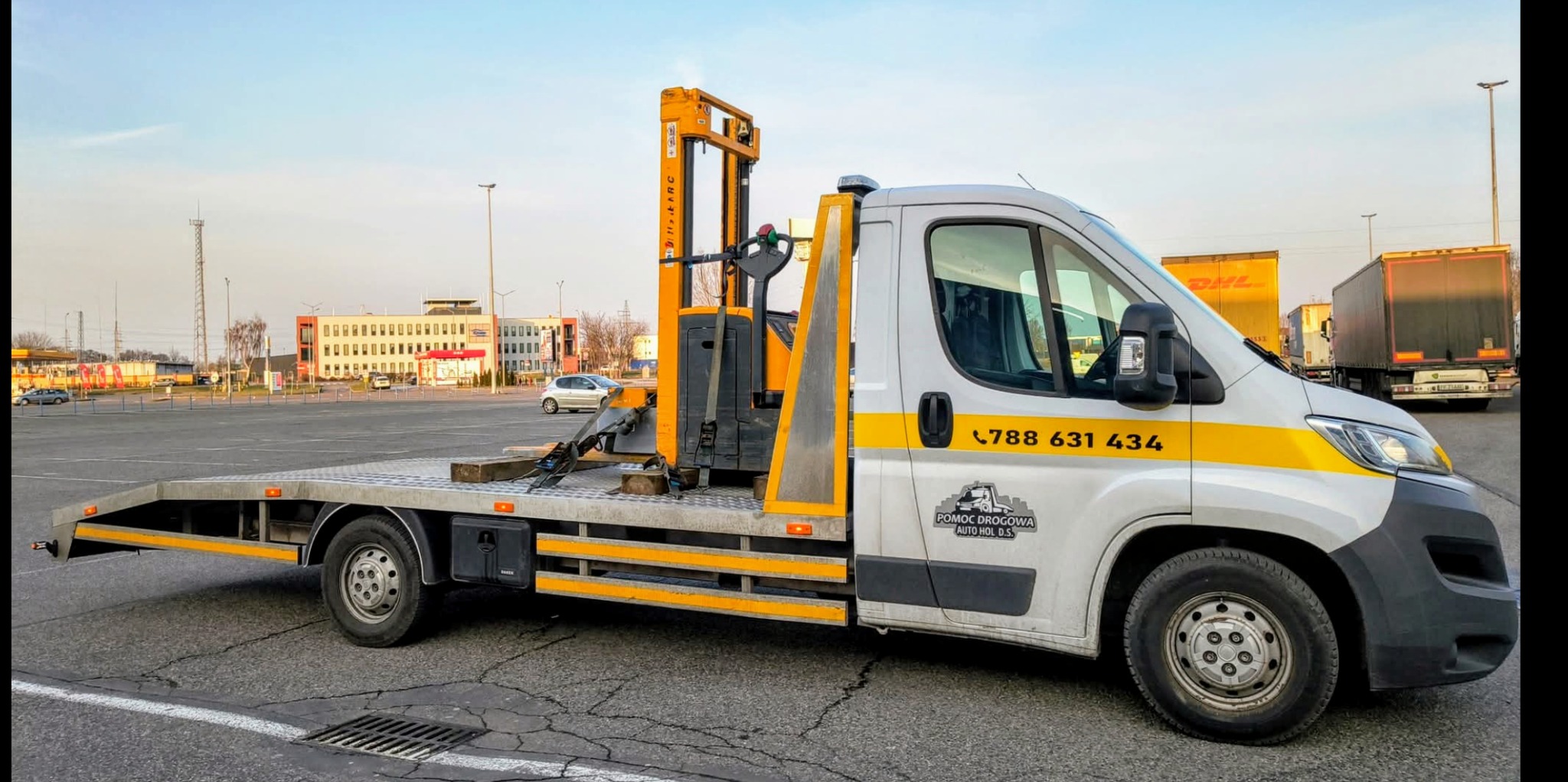 Laweta z żółtym wózkiem widłowym na platformie, zaparkowana na parkingu. Widoczny numer telefonu i logo firmy Pomoc Drogowa Auto Hol D.S.