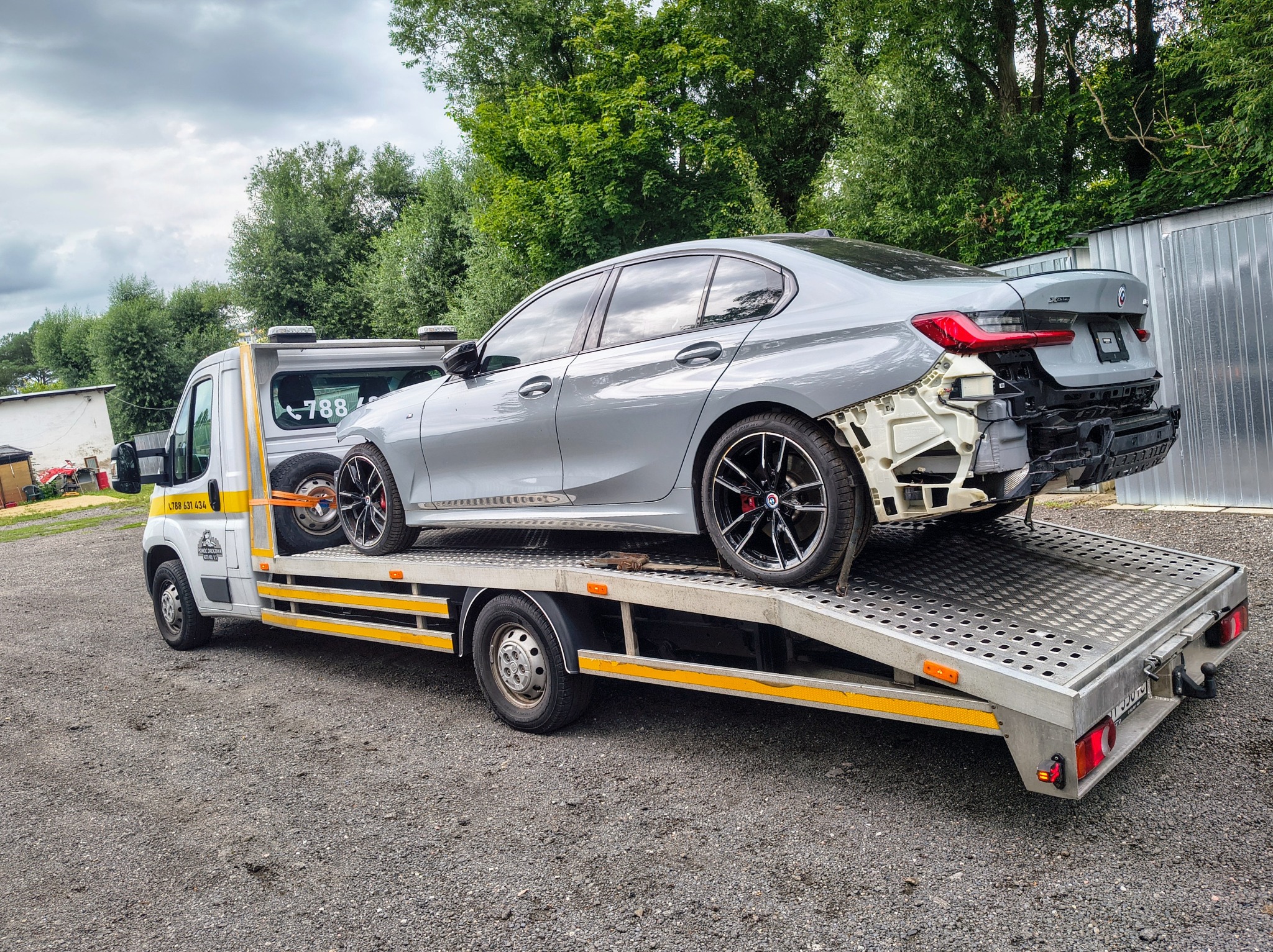 Laweta z uszkodzonym, szarym BMW serii 3 na platformie, zaparkowana na żwirowym podłożu, z zielenią w tle. Widoczny numer telefonu na kabinie lawety.