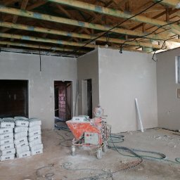 Osoba prywatna - Wnętrze w trakcie prac wykończeniowych: ściany pokryte świeżym tynkiem, widoczna maszyna tynkarska, stos worków z zaprawą i drewniana konstrukcja sufitu z widocznymi instalacjami.