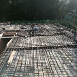 Zbrojenie stropu z siatki stalowej i betonowych podkładek dystansowych, widoczne elementy deskowania i fragment murowanej ściany z pustaków.