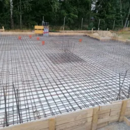 Zbrojenie płyty fundamentowej z drewnianym szalunkiem, widoczne pionowe pręty zbrojeniowe i pomarańczowe rury instalacyjne, w tle zieleń drzew.