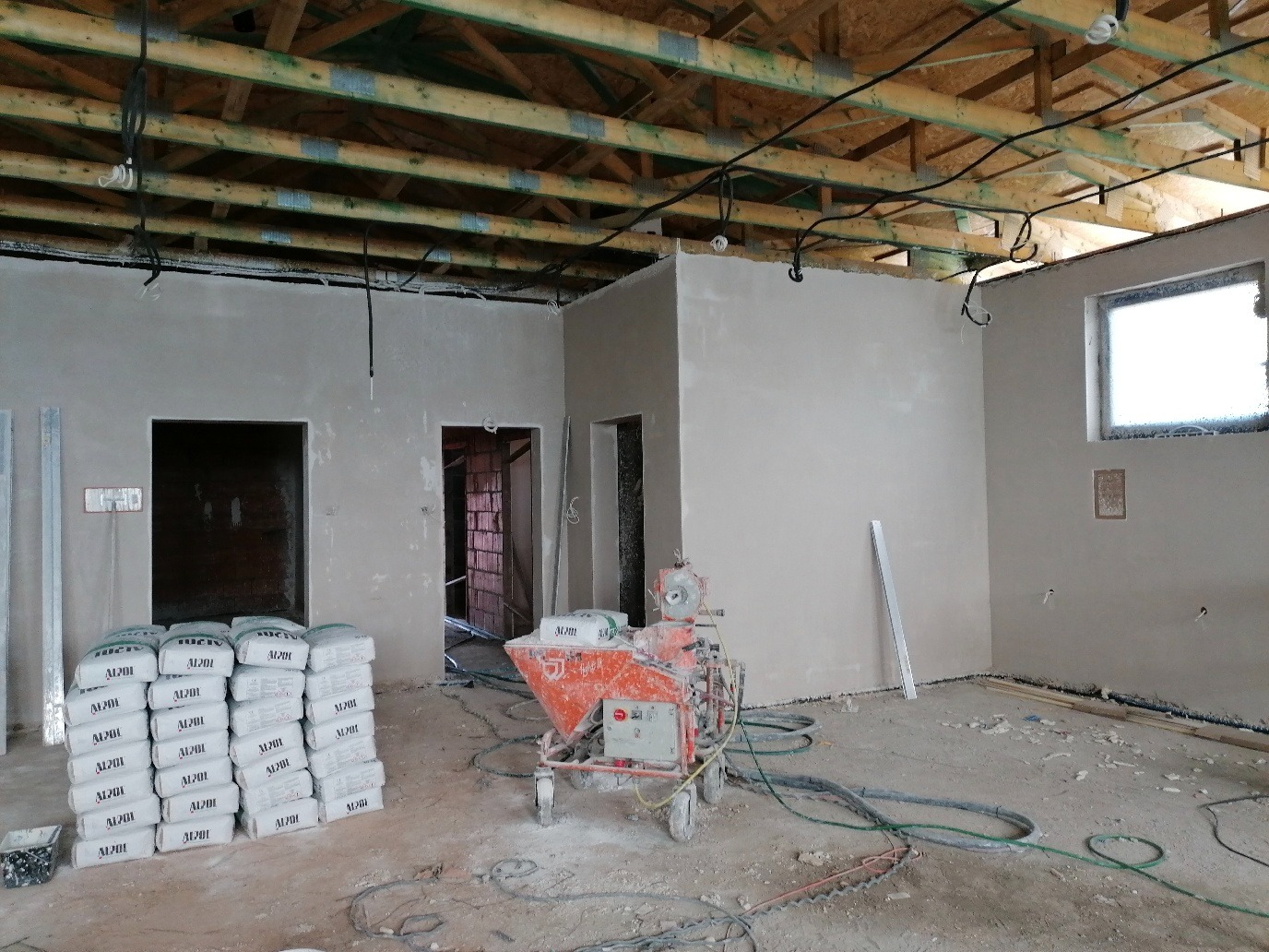 Wnętrze w trakcie prac wykończeniowych: ściany pokryte świeżym tynkiem, widoczna maszyna tynkarska, stos worków z zaprawą i drewniana konstrukcja sufitu z widocznymi instalacjami.