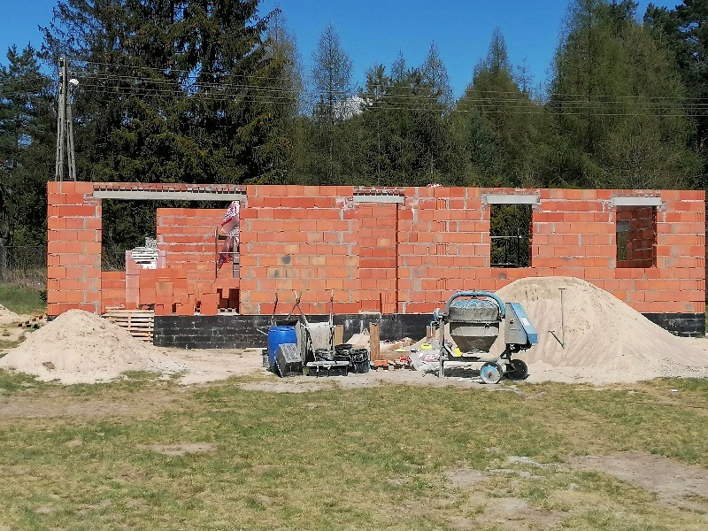Wznoszenie ścian z czerwonej cegły na budowie domu w Lutomiersku. Widoczna betoniarka, piasek i narzędzia murarskie.