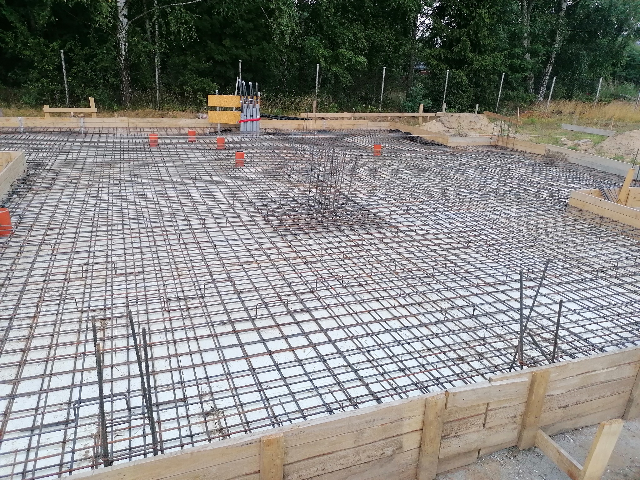 Zbrojenie płyty fundamentowej z drewnianym szalunkiem, widoczne pionowe pręty zbrojeniowe i pomarańczowe rury instalacyjne, w tle zieleń drzew.