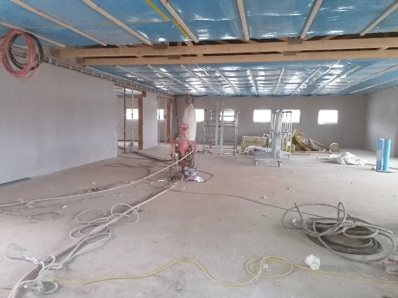 Wnętrze w trakcie prac wykończeniowych: ściany pokryte świeżym tynkiem, na podłodze rozłożone kable, maszyna do tynkowania w centralnej części pomieszczenia, sufit zabezpieczony folią, okna w tle.