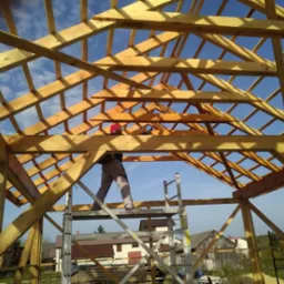 Konstrukcja drewniana dachu w trakcie budowy, widok od wewnątrz, pracownik w czerwonym kasku na aluminiowej drabinie, błękitne niebo w tle.