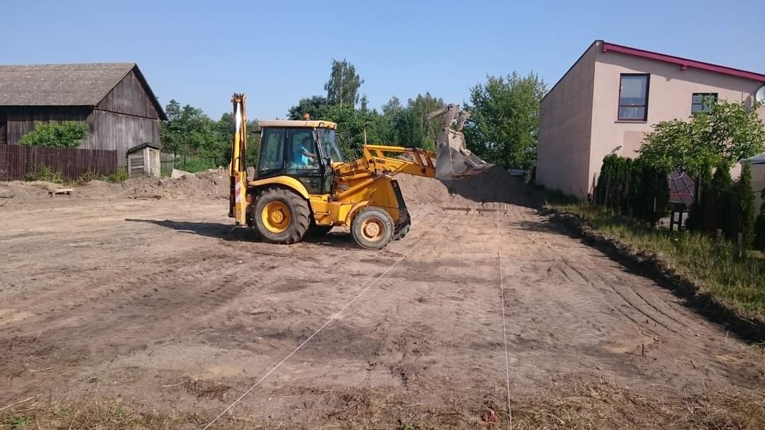 Żółta koparko-ładowarka podczas wyrównywania terenu pod budowę, widoczne ślady opon na ziemi, w tle budynki mieszkalne i gospodarcze, słoneczny dzień.