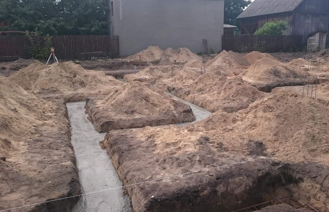 Wykopane fundamenty zalane betonem, widoczne hałdy piasku i niwelator optyczny na statywie w tle.