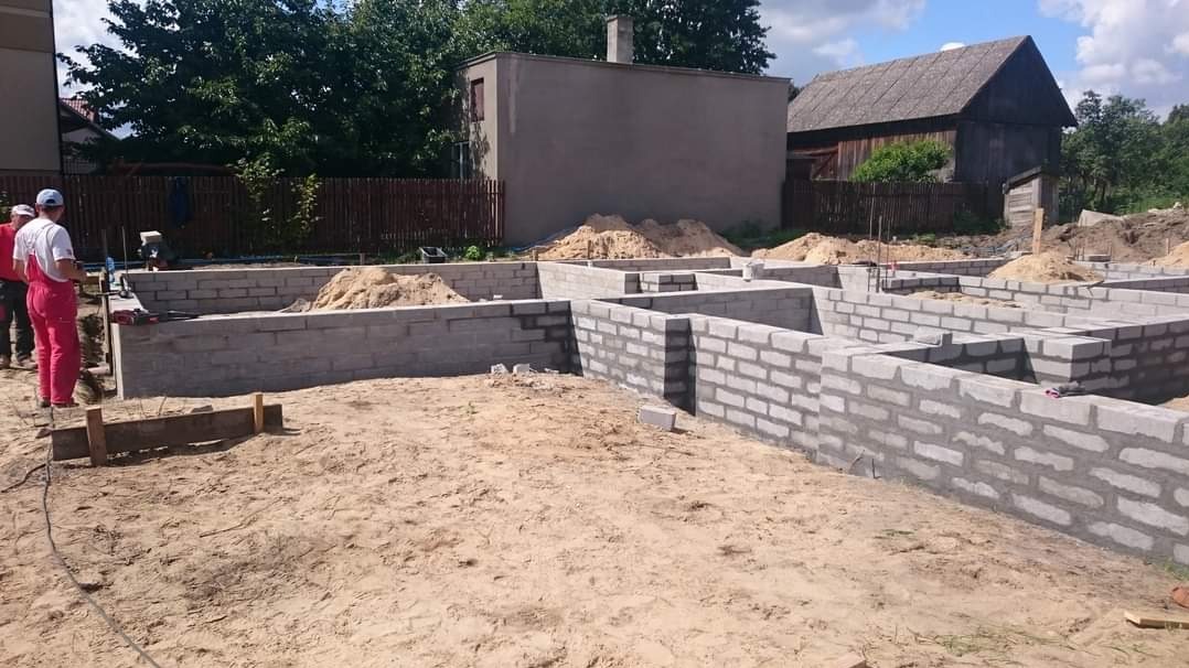 Wznoszenie fundamentów budynku z bloczków betonowych, widoczni dwaj pracownicy w roboczych ubraniach, ułożone bloczki z zaprawą, piasek w wykopach, w tle budynki mieszkalne i drzewa.