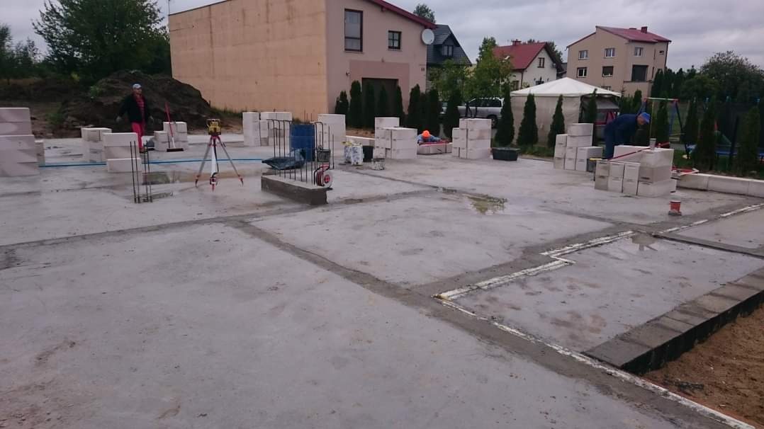 Wylany fundament z zaznaczonymi obrysami ścian, przygotowany do dalszych prac murarskich z użyciem bloczków betowych. Widoczny laser krzyżowy na statywie oraz pracownicy w tle.
