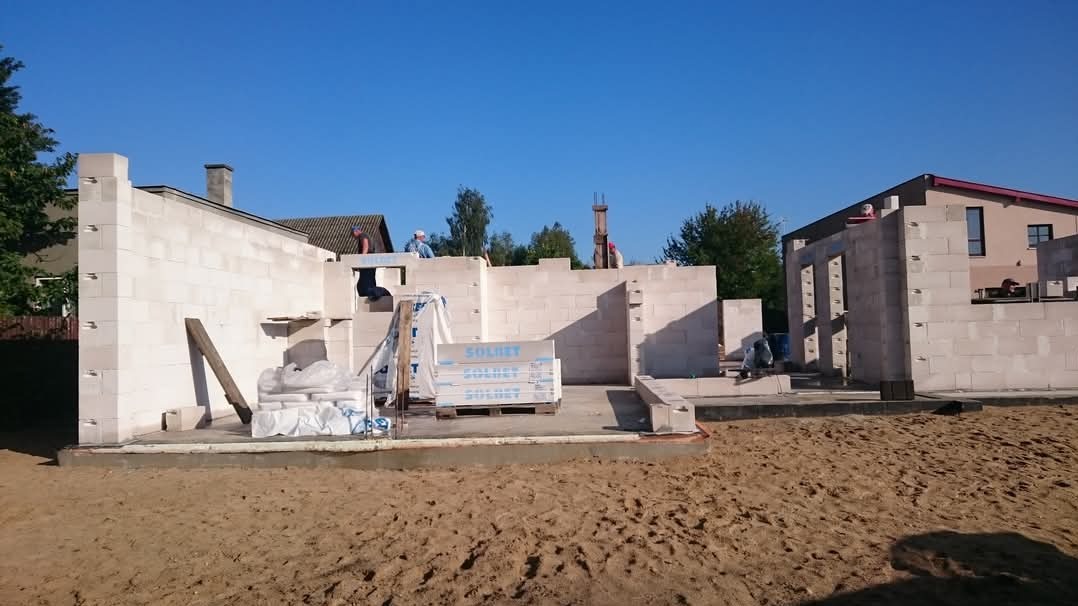 Wznoszenie ścian domu z białych bloczków Solbet, widoczni pracownicy budowlani, na pierwszym planie paleta z bloczkami i worki z zaprawą.