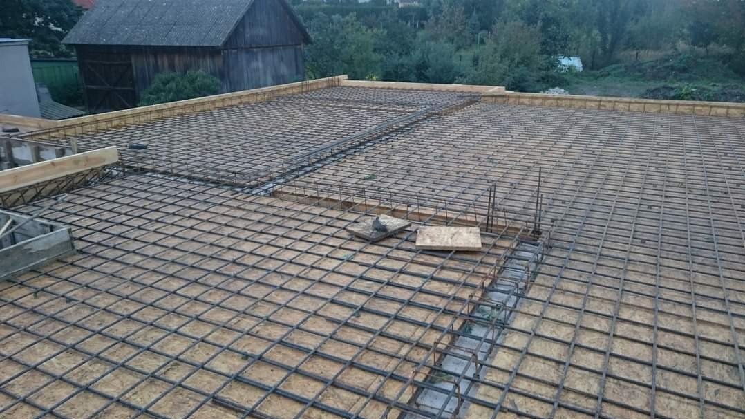 Zbrojenie stropu z siatki stalowej na deskowaniu, widoczne fragmenty szalunku i drewniane podpory, w tle budynek gospodarczy i drzewa.