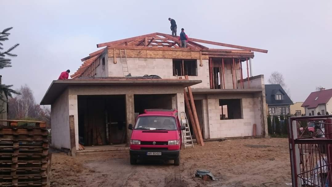 Budowa domu jednorodzinnego w stanie surowym otwartym, widoczni pracownicy na dachu, czerwony bus zaparkowany przed budynkiem, palety z materiałami budowlanymi w tle.