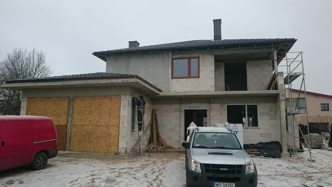 Surowy stan domu jednorodzinnego w trakcie budowy, z widoczną elewacją w stanie deweloperskim, częściowo zakrytymi oknami płytami OSB, pracownikiem na drabinie przy oknie oraz zaparkowanym...