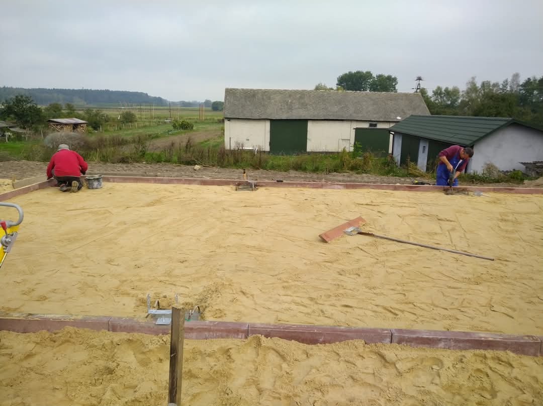 Przygotowanie podłoża pod budowę tarasu z obrzeżami z kostki, widoczni dwaj pracownicy wyrównujący piasek, w tle budynki gospodarcze.