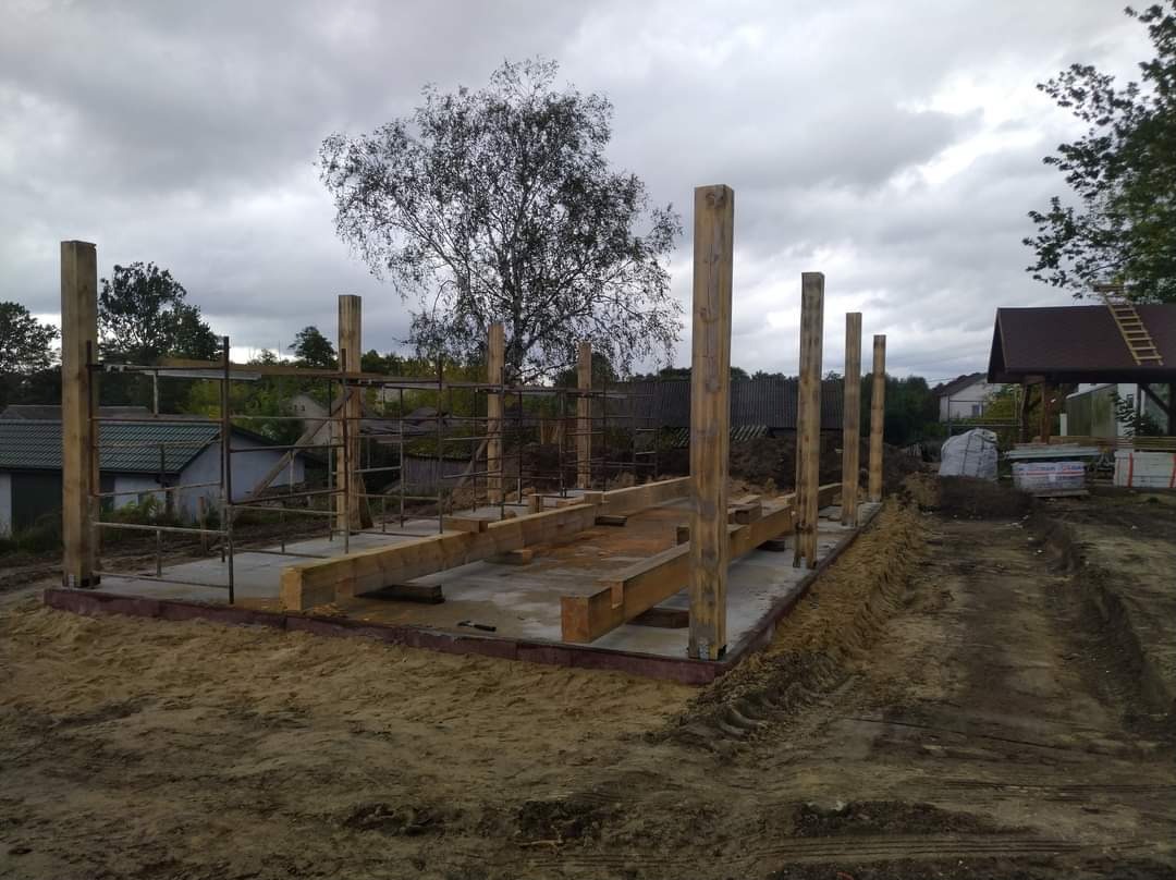 Surowy szkielet drewnianej konstrukcji na betonowej podstawie, z widocznymi słupami i belkami, otoczony ziemią i w tle z zabudowaniami mieszkalnymi.