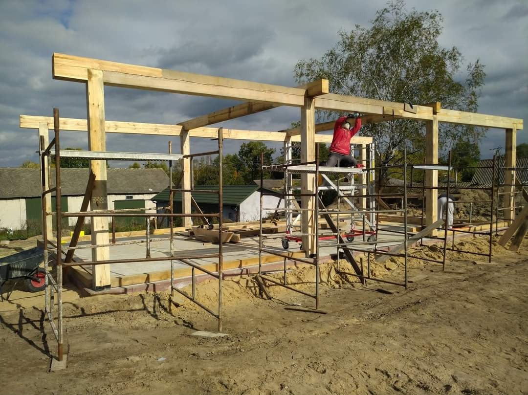 Konstrukcja szkieletowa budynku w trakcie budowy, widoczny pracownik na rusztowaniu w czerwonej bluzie, w tle zabudowania gospodarcze i drzewa.