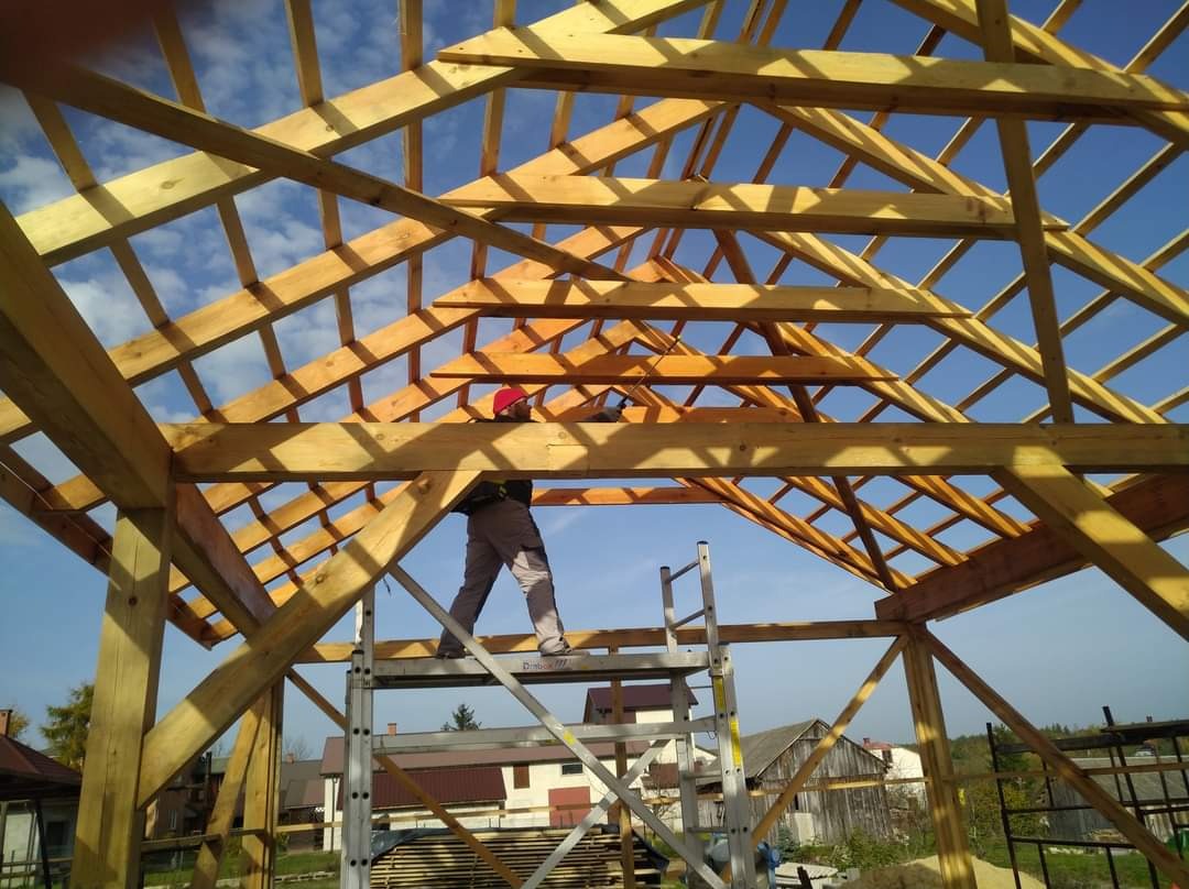 Konstrukcja drewniana dachu w trakcie budowy, widok od wewnątrz, pracownik w czerwonym kasku na aluminiowej drabinie, błękitne niebo w tle.