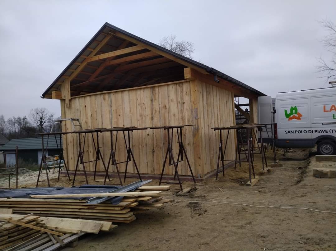 Wznoszenie drewnianej konstrukcji szkieletowej budynku z widocznym dachem, wspartej na metalowych kozłach, z deskami ułożonymi na ziemi i samochodem dostawczym w tle.