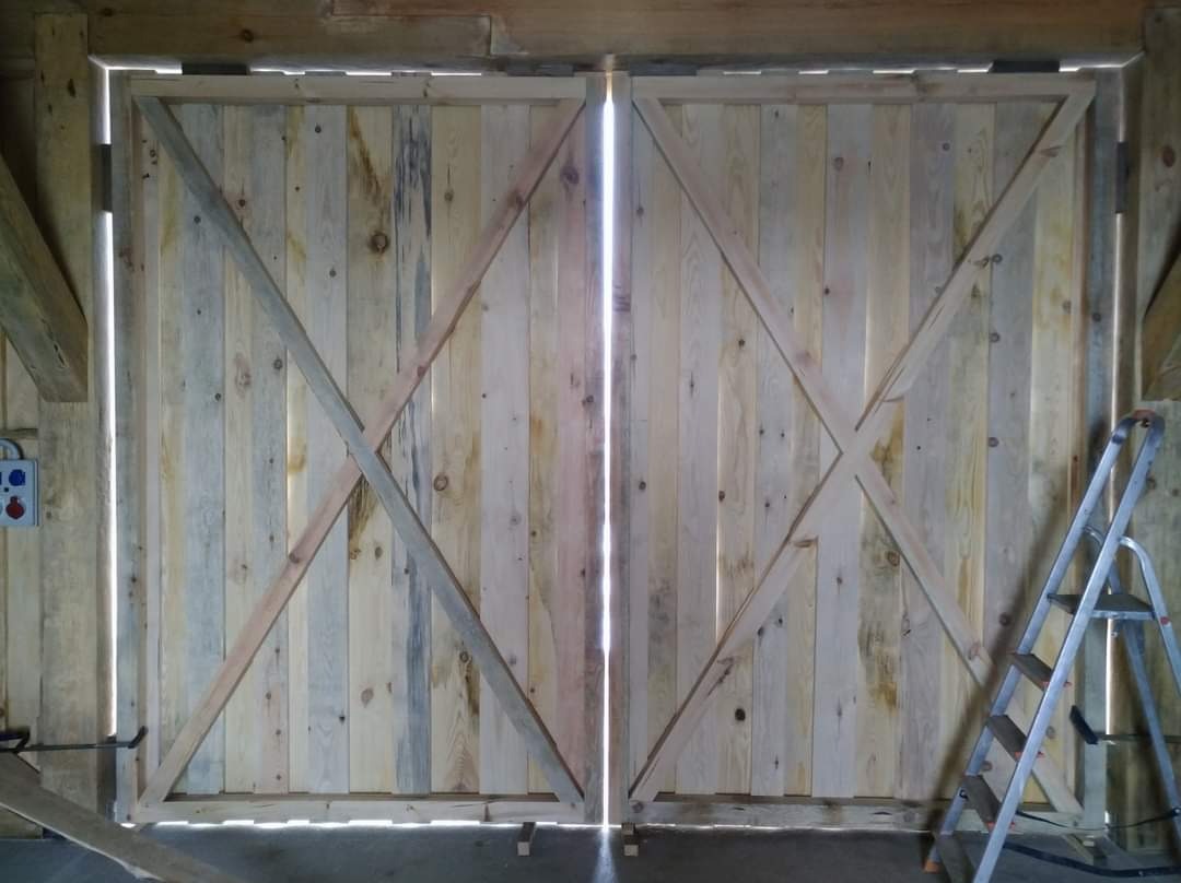 Dwuskrzydłowe drzwi stodoły wykonane z surowych, pionowo ułożonych desek z ukośnymi wzmocnieniami, widoczne szczeliny między deskami przepuszczające światło, drabina przystawiona z prawej strony.
