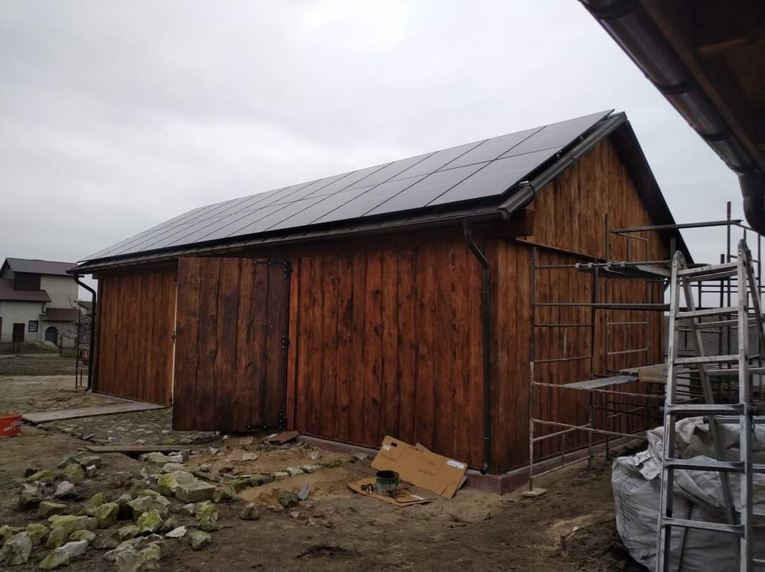 Drewniany budynek gospodarczy z panelami fotowoltaicznymi na dachu, z częściowo otwartymi wrotami i rusztowaniem z boku, w trakcie prac wykończeniowych na zewnątrz.