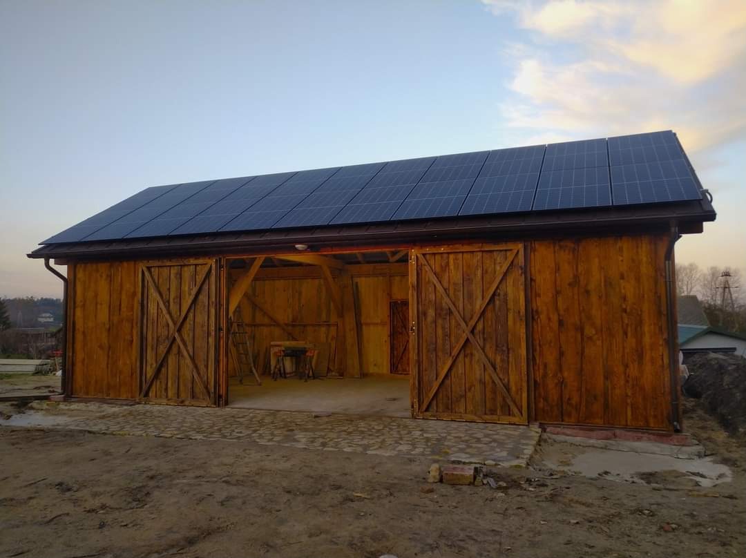 Drewniany budynek gospodarczy z otwartymi wrotami i zamontowanymi panelami słonecznymi na dachu, widoczne wnętrze z drabiną i stołem roboczym, teren wokół budynku częściowo utwardzony kostką.