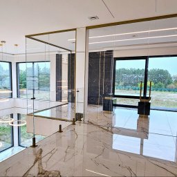 CNL Tomasz Pietrucha - Nowoczesne wnętrze z szklaną balustradą na antresoli, marmurowa podłoga z widocznymi odbiciami, duże okna z widokiem na zieleń i designerskie lampy wiszące.