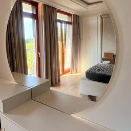 CNL Tomasz Pietrucha - Okrągłe lustro w łazience odbija fragment pokoju z oknem i zasłonami, widoczne białe meble łazienkowe z złotymi uchwytami.