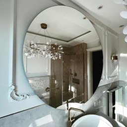 CNL Tomasz Pietrucha - Owalne lustro w eleganckiej łazience odbija kabinę prysznicową z marmurowymi ścianami i złotą armaturą, a także designerski żyrandol w kształcie gałęzi z białymi listkami.