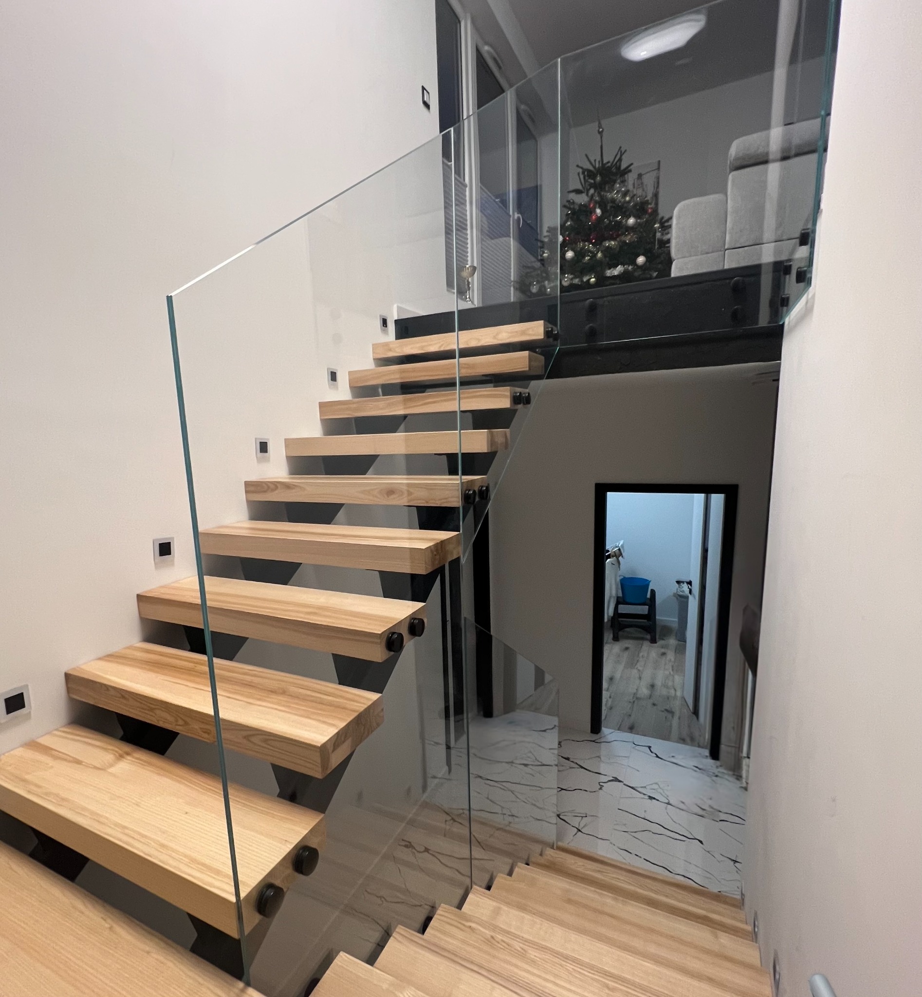 Nowoczesne schody drewniane z balustradą szklaną, mocowane punktowo, z widocznym fragmentem świątecznej choinki w tle.