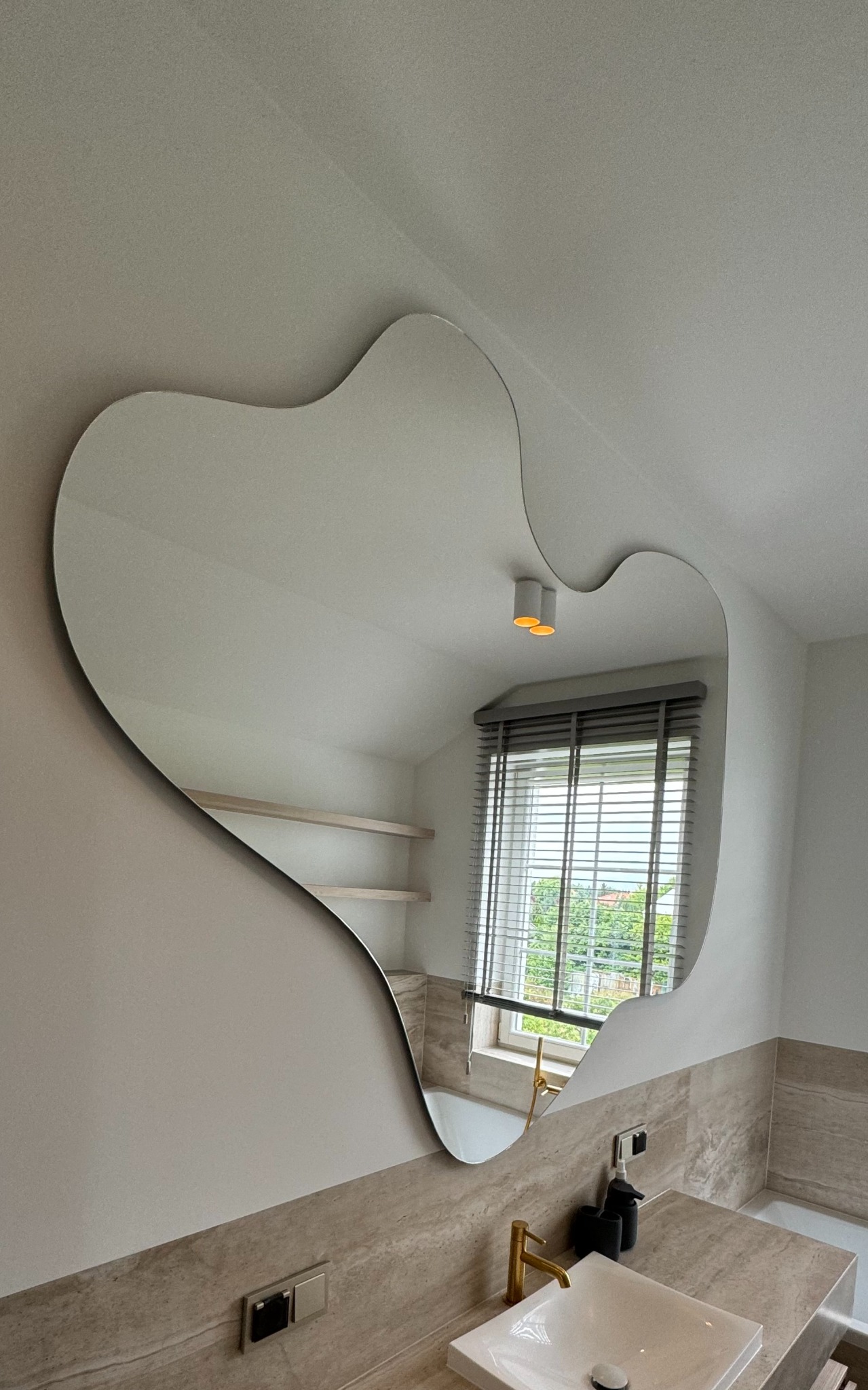 Nieregularne, ozdobne lustro na ścianie w łazience z beżowymi płytkami, odbijające okno z żaluzjami i fragment sufitu z lampą.