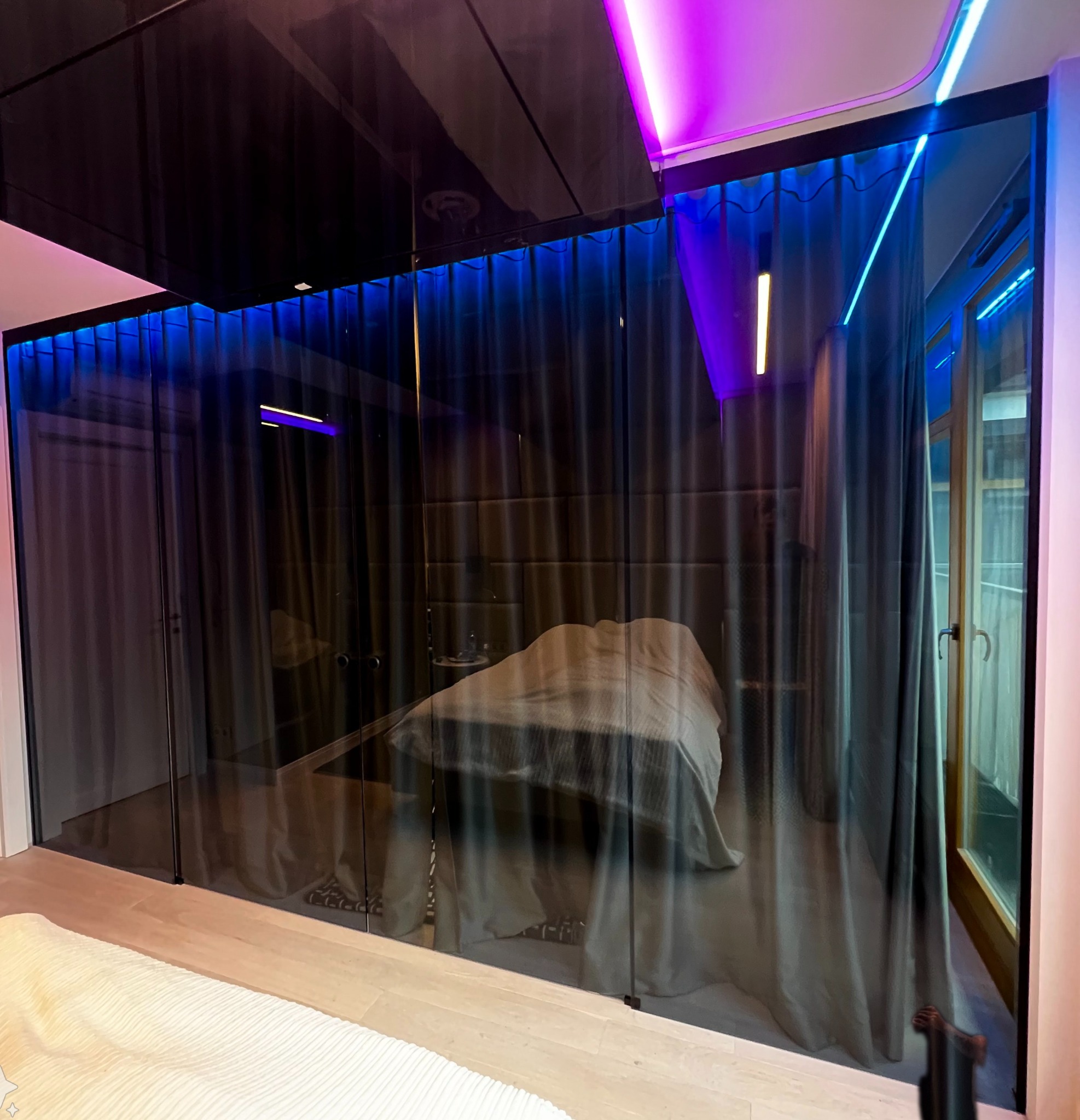 Szklana ściana działowa oddzielająca sypialnię, podświetlona niebieskim i fioletowym światłem LED, widok na łóżko z szarą pościelą i zasłony.