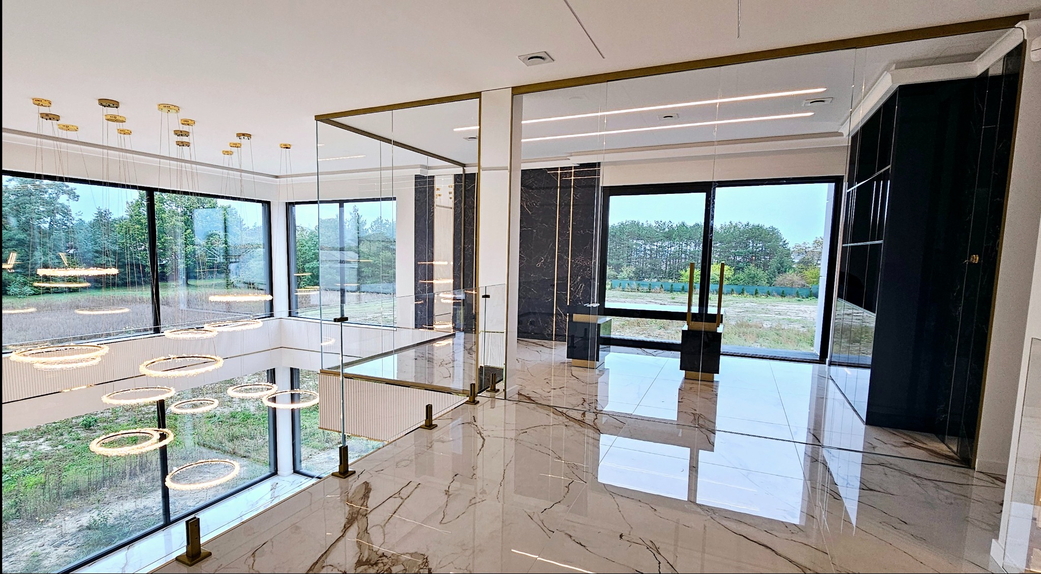 Nowoczesne wnętrze z szklaną balustradą na antresoli, marmurowa podłoga z widocznymi odbiciami, duże okna z widokiem na zieleń i designerskie lampy wiszące.