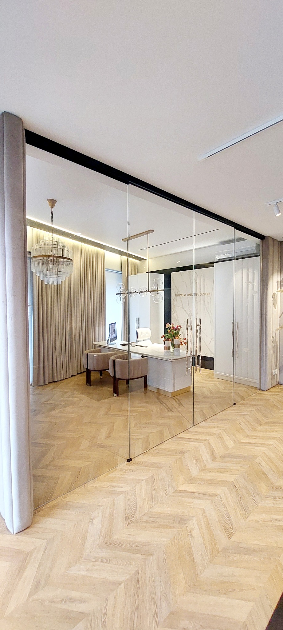 Przeszklone biuro w Zalesiu Górnym z widokiem na eleganckie wnętrze z parkietem w jodełkę, designerskim biurkiem i krzesłami oraz dekoracyjnym żyrandolem.