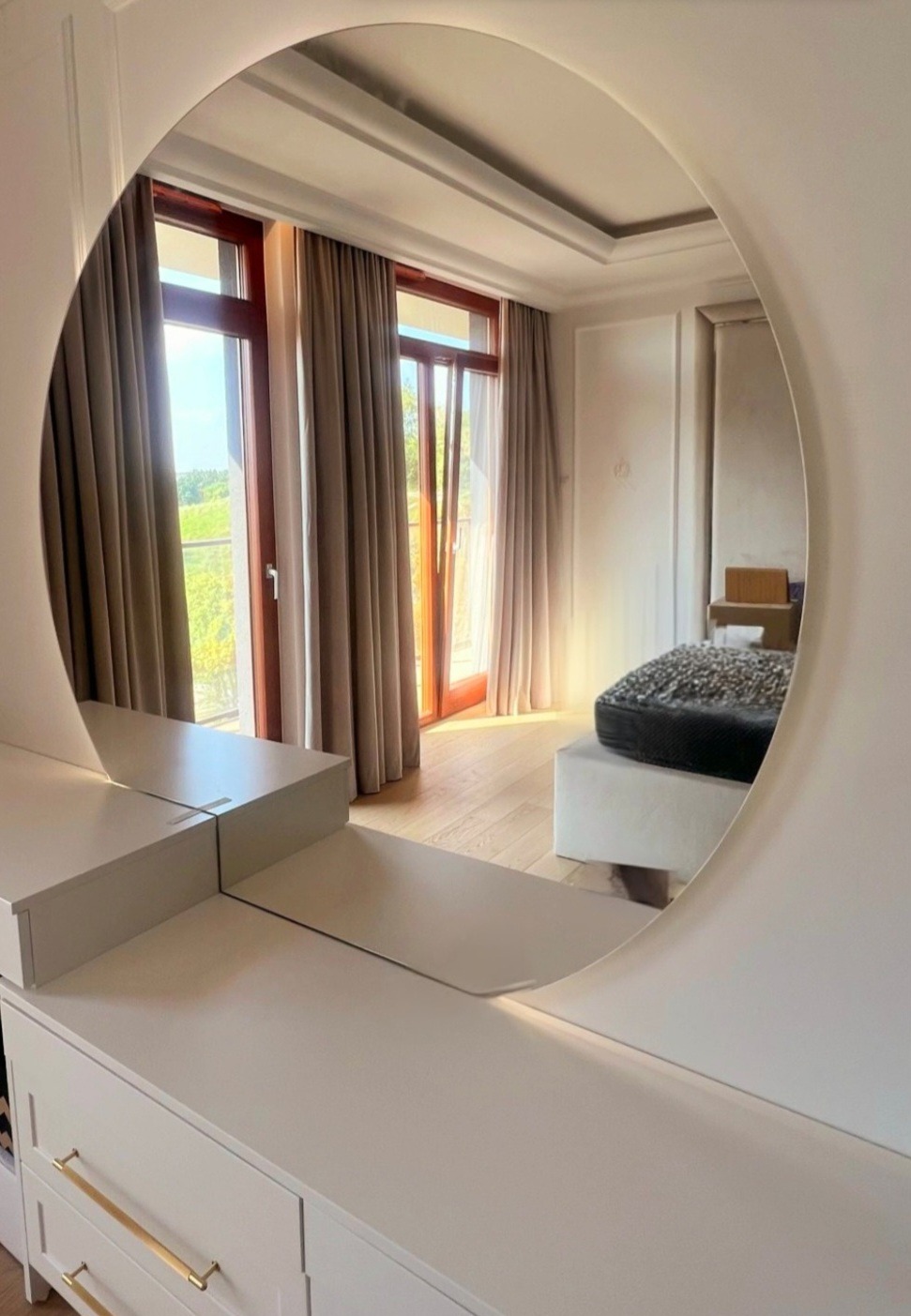 Okrągłe lustro w łazience odbija fragment pokoju z oknem i zasłonami, widoczne białe meble łazienkowe z złotymi uchwytami.