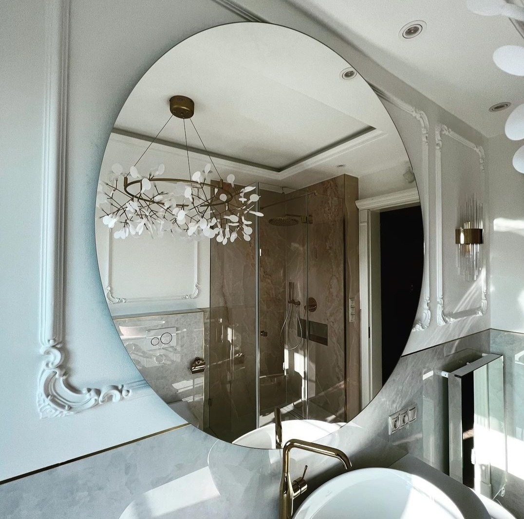 Owalne lustro w eleganckiej łazience odbija kabinę prysznicową z marmurowymi ścianami i złotą armaturą, a także designerski żyrandol w kształcie gałęzi z białymi listkami.