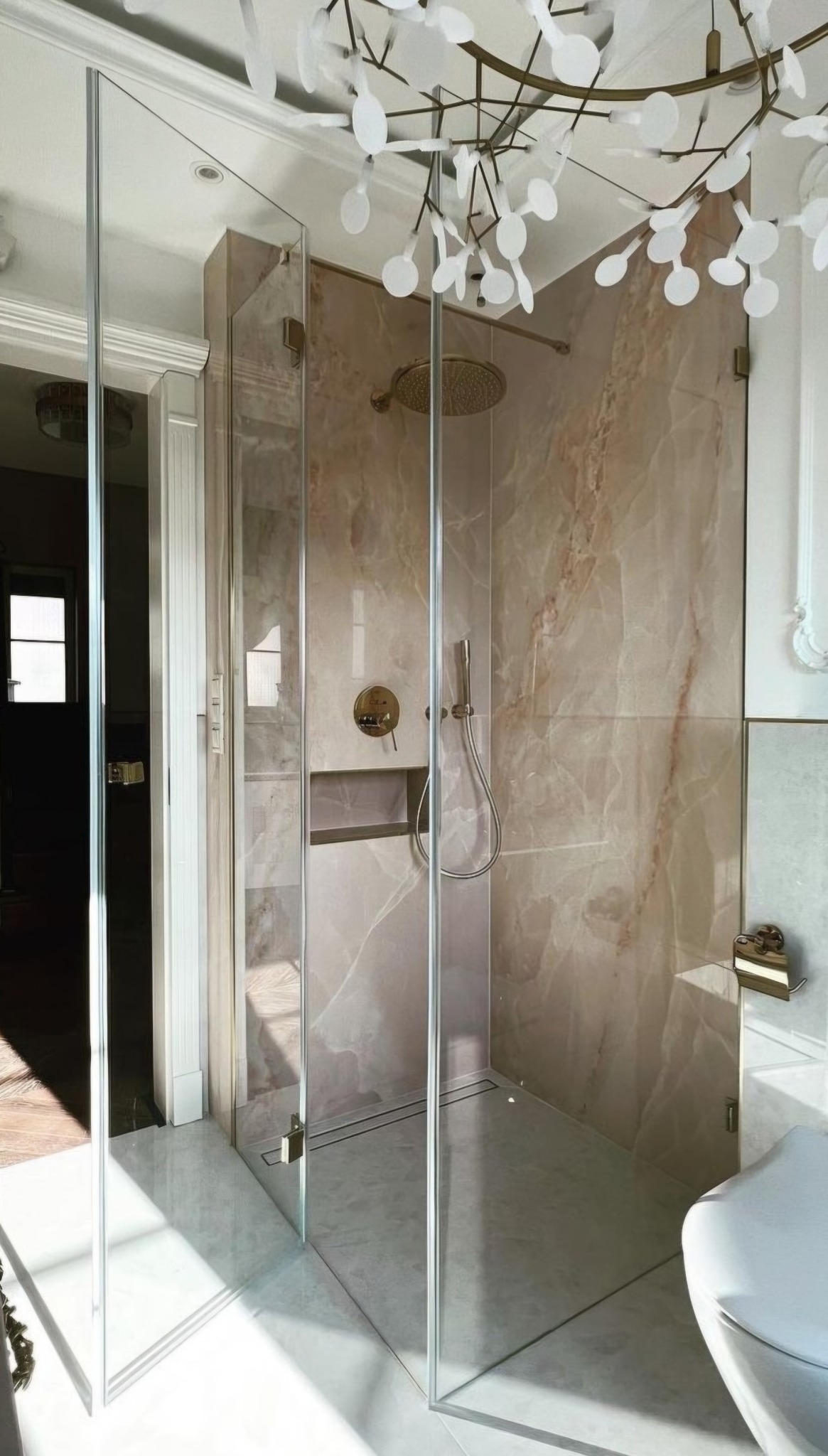 Nowoczesna kabina prysznicowa ze szklanymi drzwiami, wykończona marmurem w odcieniach beżu i złota, oświetlona dekoracyjnym żyrandolem w kształcie gałęzi z białymi listkami.